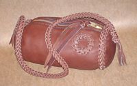  This braided Mahogany bag has a large brass zipper and is about 9" long by a 5" diameter. It has an 8 strand round braided shoulder strap with short tassels on the ends of it. There is a braided circle applique on the side of it. 